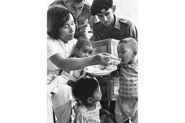 This black-and-white photograph shows two Australian soldiers from the 1st Australian Task Force providing civilian food aid to a Vietnamese woman and her three children in Phuoc Tuy province during the Vietnam War